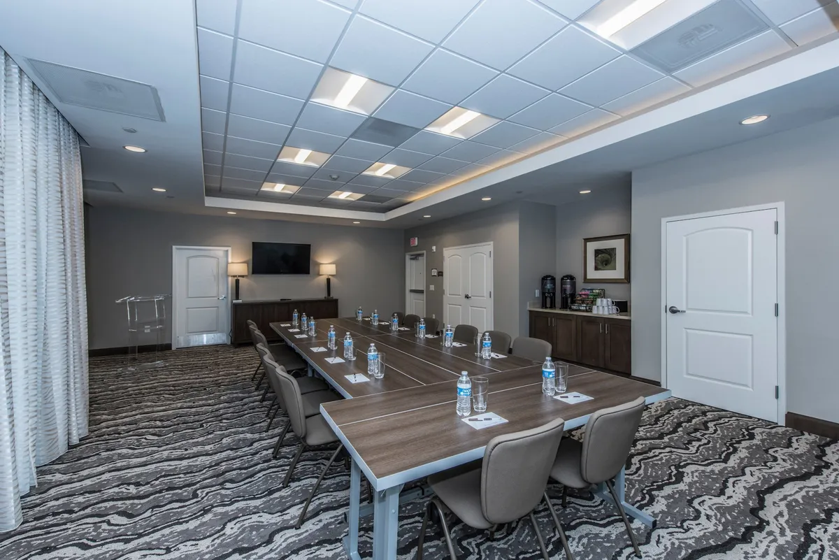 A modern conference room featuring a long conference table set with water bottles, a television screen for presentations, and ergonomic chairs. The neutral décor and ample table space create an environment ideal for meetings, training sessions, or collaborative work.