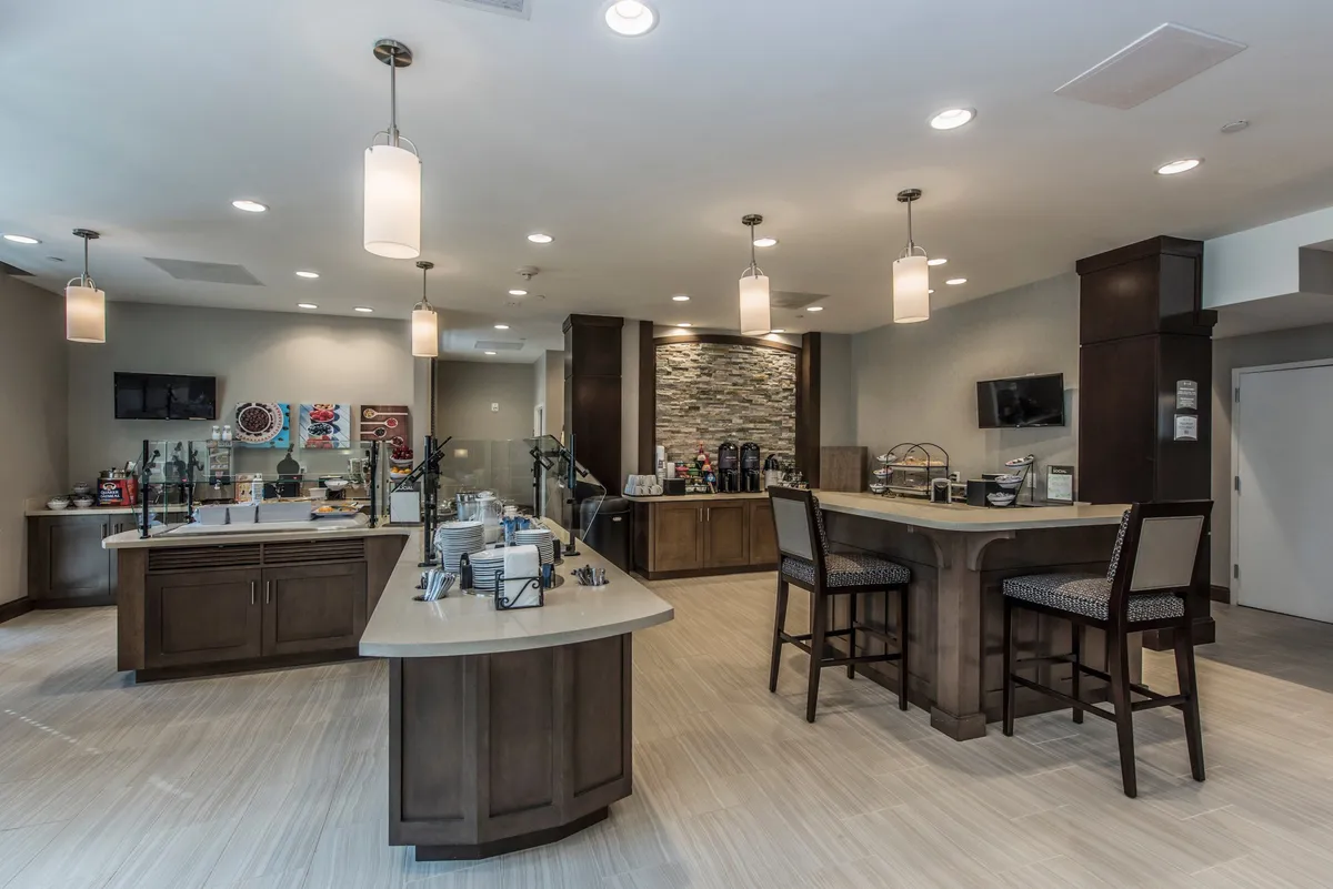 The image displays a cozy breakfast bar area with a stone accent wall, a central island featuring seating, and a well-stocked beverage station. Natural light enhances the inviting atmosphere, making it a perfect spot for guests to gather and enjoy the complimentary breakfast buffet or coffee throughout the morning.