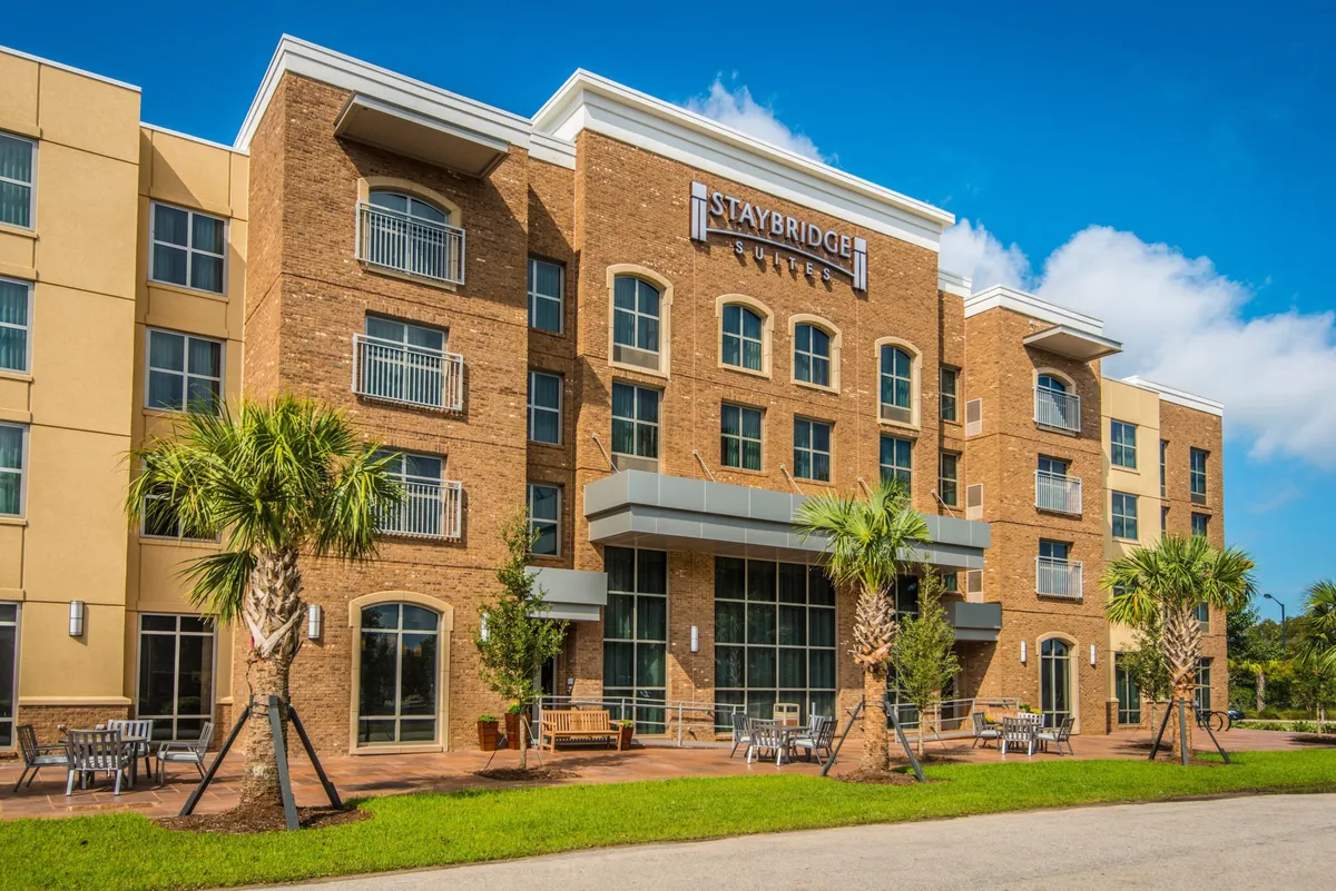 This image showcases the main entrance of Staybridge Suites Mount Pleasant, framed by large windows and surrounded by landscaped palm trees and inviting outdoor seating. The Staybridge Suites sign is clearly visible, conveying a welcoming first impression for guests arriving for business or leisure stays.