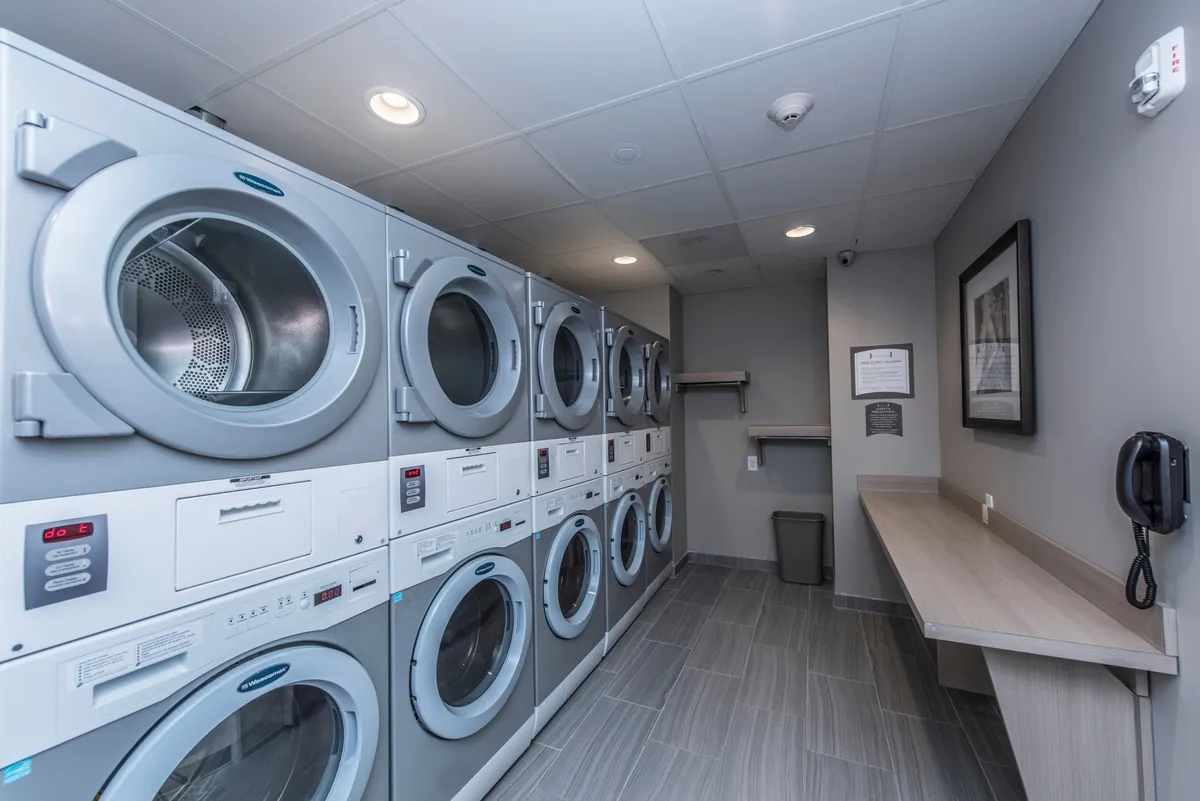 A practical laundry room equipped with laundry machines, a countertop surface, complementing wall art, and informational signage. The setup is organized and designed for guest convenience during longer stays.