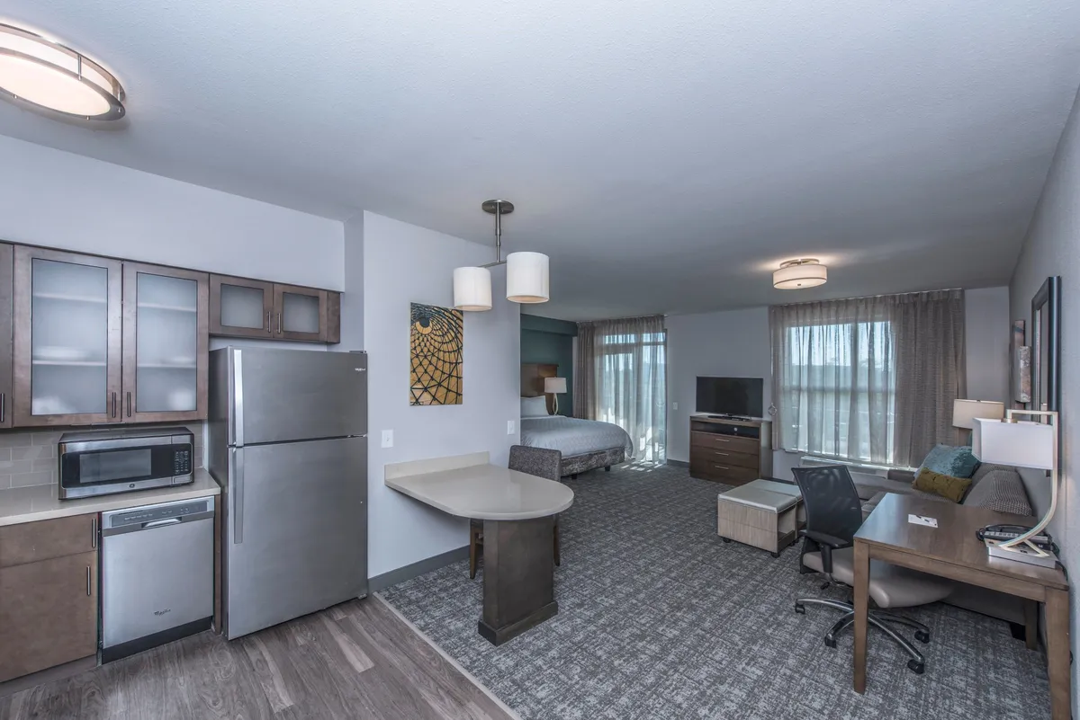 The image showcases a stylish, well-lit studio interior featuring a cozy bed, modern kitchenette, inviting dining table, sleek desk, and a mounted television. The arrangement is open and thoughtfully designed to support relaxation, cooking, working, and unwinding, all within a comfortable, contemporary space.