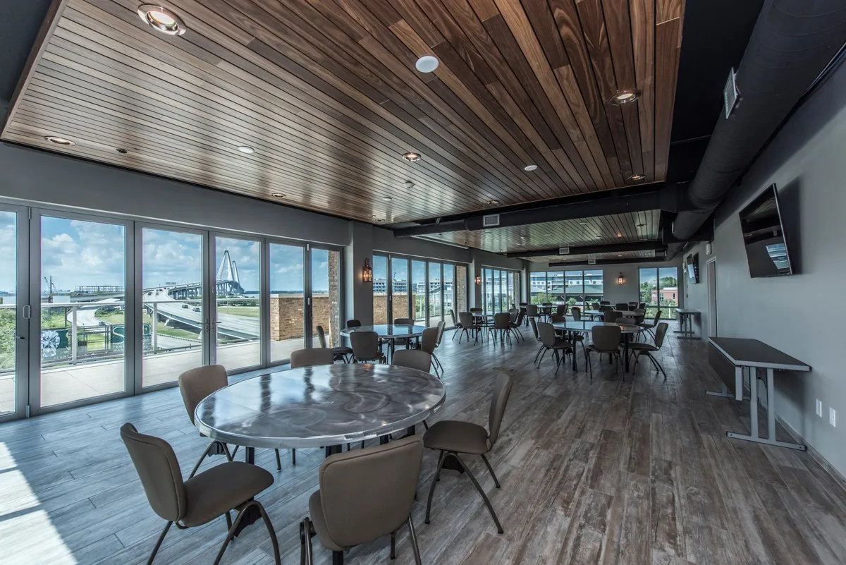 A spacious interior common area with large windows letting in natural light, round tables ideal for informal meetings, and a modern wooden ceiling. The design is open and contemporary, supporting both social gatherings and business functions.