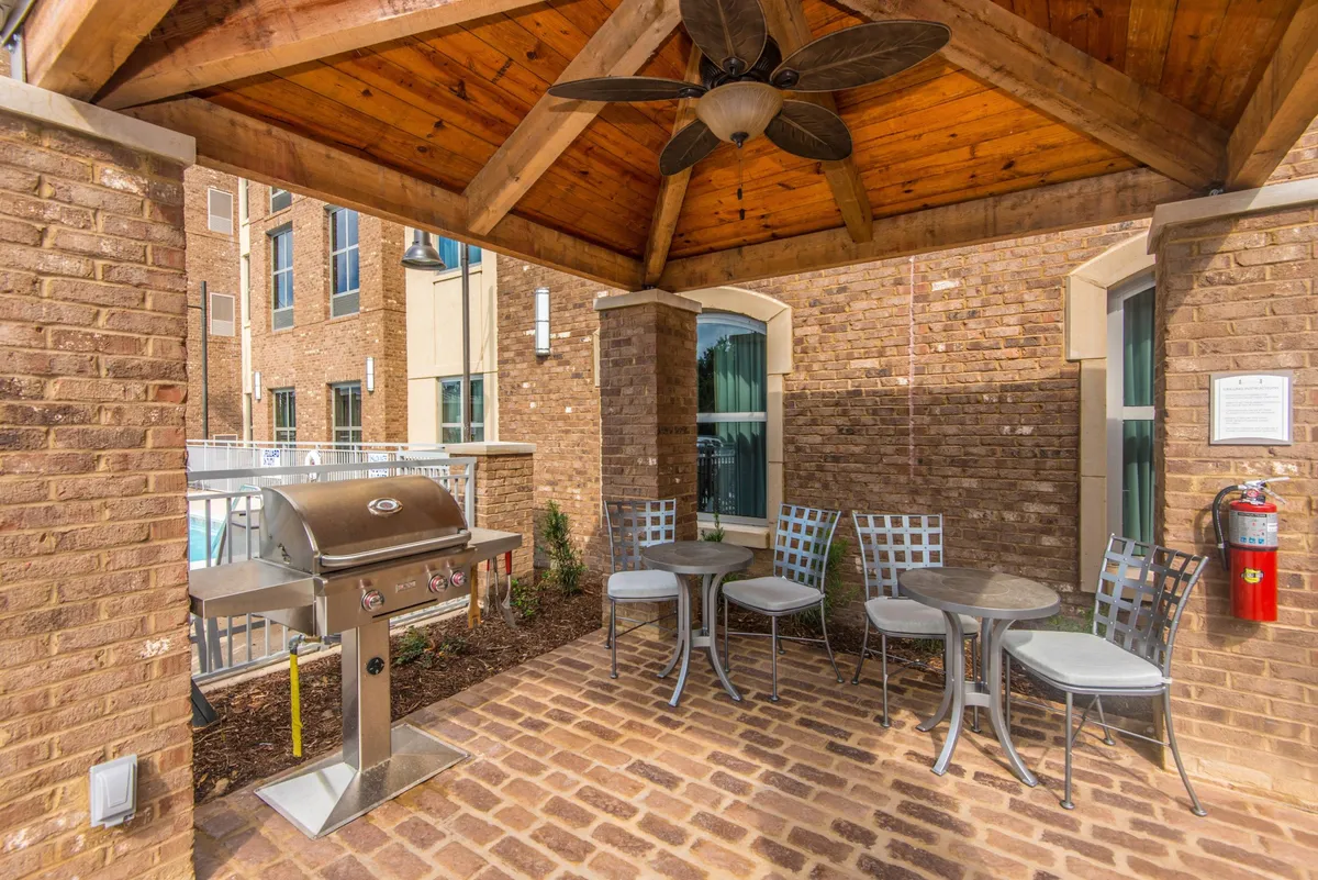 A welcoming outdoor seating area with a wooden ceiling and brick walls, featuring a communal grill and comfortable spots for guests to relax. Groups of people are seen socializing and enjoying a barbecue, creating a lively, festive atmosphere reminiscent of summer gatherings.